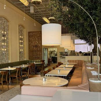 La Sorella - Decor - The photo depicts the interior of La Sorella restaurant, with a mix of seating options including a long pink banquette, round and rectangular tables, and wooden chairs. Large white cylindrical pendant lamps hang from the ceiling, which has an exposed grid structure with additional lighting fixtures. A tall green plant is visible on the right side, and the background includes a view of the kitchen area and menu display. The decor combines modern and classic elements, creating a spacious dining environment. - Breakfast & Brunch Near Me - Fishkill, New York
