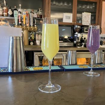 Le Sel - Mimosa - Two mimosas in tall, narrow glasses are positioned on a bar counter, one with a yellow hue and the other with a purple hue. The bar area features bar tools, metal cups, and bottles of alcohol in the background, along with wooden cabinetry and a small monitor. The setting appears to be a bar or restaurant environment. - French Near Me - Washington, DC, District of Columbia