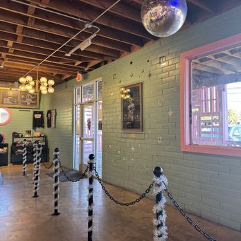 Voodoo Doughnut - The interior features a pastel green brick wall with a window and framed artwork. A large disco ball hangs from the exposed wooden ceiling. Decorative poles with black tops, chains, and white tinsel are arranged in a line, creating a barrier. The space includes modern lighting fixtures and a menu board, indicating a retail or cafe setting. - Donuts Near Me - Austin, Texas