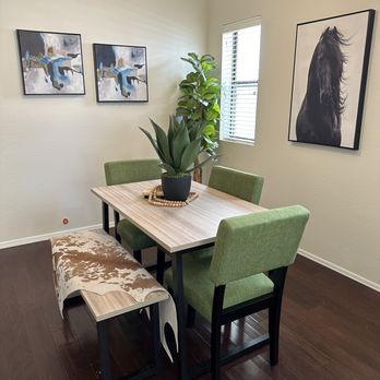 Pearson Moving - A dining room features a wooden table with green upholstered chairs, a bench with a cowhide pattern, and wall art including a horse portrait and abstract paintings. A potted plant is on the table near a window. - Movers Near Me - Gilbert, Arizona