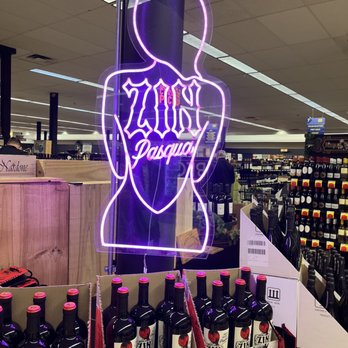 The Party Source - A neon sign in purple outlines a stylized female figure with a halo, displaying "Zaino Pasqua" in a liquor store aisle with wine bottles. - Tobacco Shops Near Me - Bellevue, Kentucky