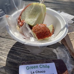 a paper plate of food and a sign that reads green chile - A foam cup holds a slice of apple and a piece of fried food, with a toothpick inserted into the apple slice. The cup is on a weathered wooden surface outdoors. A packaged baked good labeled "Green Chile" from La Choco in Albuquerque is positioned in front of the cup. The scene suggests a casual outdoor food setting, possibly at a market or food stand.