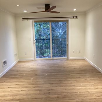 Baurer Construction & Flooring - Living Room - The image shows an empty living room with light-colored hardwood flooring and white walls. A ceiling fan is installed in the center of the ceiling, which also has recessed lighting. A sliding glass door with a curtain rod above it leads to a balcony, revealing a view of trees outside. The room has multiple electrical outlets and a vent on the lower part of one wall. - Flooring Near Me - Gresham, Oregon