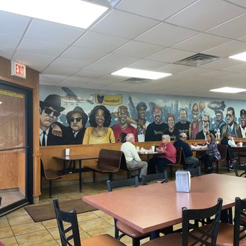 Herm's Palace - Inside Herms Palace - The image shows the interior of Herm's Palace, a family-run diner, with a mural on the wall depicting diverse individuals, including a woman in yellow and men in suits and casual attire. Several customers are seated at tables, eating and talking. The ceiling has fluorescent lighting and air vents, and the floor is tiled. The restaurant has a casual atmosphere with indoor seating. - American Near Me - Skokie, Illinois