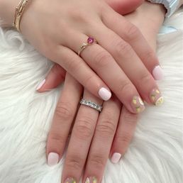 Lemon design - The image shows two hands with manicured nails, painted in light pink with lemon slice designs on some nails. The hands are adorned with rings and bracelets, and one wrist has a white digital watch. The hands are resting on a soft, white furry surface, and the photo captures details of jewelry and nail art.