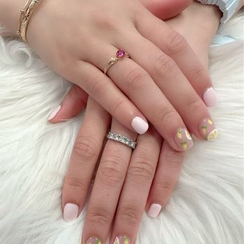 Solar Lashes & Nails Spa - Lemon design - The image shows two hands with manicured nails, painted in light pink with lemon slice designs on some nails. The hands are adorned with rings and bracelets, and one wrist has a white digital watch. The hands are resting on a soft, white furry surface, and the photo captures details of jewelry and nail art. - Eyelash Service Near Me - Boston, Massachusetts