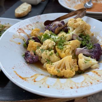 Debajo Tapas ABQ - Roasted Cauliflower - A white round plate holds roasted cauliflower florets garnished with chopped herbs, accompanied by purple cauliflower pieces. The dish is presented with a light sauce or oil, and a pair of tongs rests on the plate. In the background, there are other dishes and a piece of bread on a dark table surface. - Spanish Near Me - Albuquerque, New Mexico