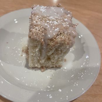 Mo's Egg House - Coffee cake - A square slice of coffee cake with a crumb topping and icing on top is placed on a white plate. The cake appears moist and crumbly, with some crumbs scattered on the plate. The background shows a wooden table surface, indicating a casual dining setting. - Breakfast & Brunch Near Me - Temecula, California