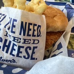 Butterburger Cheese Value Baskets - A fried fish sandwich features a golden-brown breaded fish fillet inside a soft bun, accompanied by a side of crinkle-cut fries and a pickle slice. The sandwich is partially wrapped in branded paper, and the tray includes additional pickles and condiments. The setting appears to be a casual dining or fast-food environment with blue trays and paper wrappers.