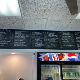 Menu - A black chalkboard menu lists various food items including gyros, hot dogs, chicken sandwiches, and side orders. The menu includes prices and descriptions for each item, such as gyro plates, grilled chicken sandwiches, and kids' menu options. The ceiling above features fluorescent lighting, and part of a refrigerator with beverages is visible below the menu.