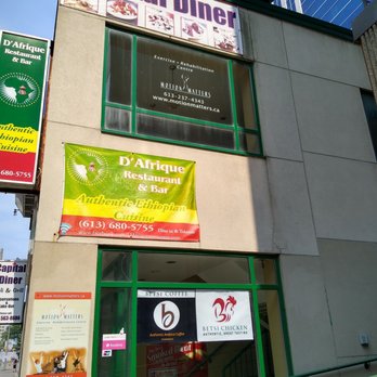 Betsi Coffee - Side of the restaurant. Besti Chicken is inside D'Afrique Restaurant and Bar. - The photo captures the exterior of a commercial building with multiple signs. The prominent signs include D'Africa Restaurant & Bar, advertising authentic Ethiopian cuisine, and Betsi Coffee, which offers Arabic coffee. Additional signage for businesses like Motion Matters and Capital Diner is visible. The building features large windows with green framing, and the street scene includes a cityscape with other buildings and traffic in the background. - Coffee Roasteries Near Me - Ottawa, Ontario