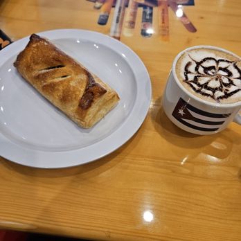 Chichos - Pastelito y cortadito - A pastry on a white plate and a mug of coffee with latte art are placed on a wooden table, with a menu or flyers in the background. - Bakeries Near Me - Pflugerville, Texas