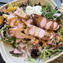 Beet & Ricotta Salad - A salad featuring mixed greens, bread croutons, diced yellow fruit, dried berries, and a pink dressing. The salad is served in a disposable bowl on a speckled countertop, with some wires visible in the background.