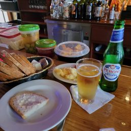 Quite the way to start! - The image shows a bar counter with a partially eaten piece of bread on a white plate, a glass of beer, and a green Peroni beer bottle. There are containers of olives, cheese, and spreads on the counter. In the background, there are shelves with various bottles of alcohol, including wine and spirits, and a small refrigerator. The setting appears to be a casual bar or restaurant environment.