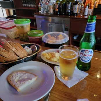 Pasquale Rigoletto Restaurant - Quite the way to start! - The image shows a bar counter with a partially eaten piece of bread on a white plate, a glass of beer, and a green Peroni beer bottle. There are containers of olives, cheese, and spreads on the counter. In the background, there are shelves with various bottles of alcohol, including wine and spirits, and a small refrigerator. The setting appears to be a casual bar or restaurant environment. - Italian Near Me - Bronx, New York