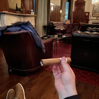 Biggs Mansion - The image shows a person seated in a vintage-style lounge with leather armchairs and sofas, holding a lit cigar in their right hand. The room has wooden paneling, curtains, and a chandelier, creating an elegant atmosphere. The person's legs, wearing tan shoes, are visible in the foreground, and a coat is draped over one of the chairs. The setting appears to be a cigar lounge or a similar upscale gathering space. - Tobacco Shops Near Me - Chicago, Illinois