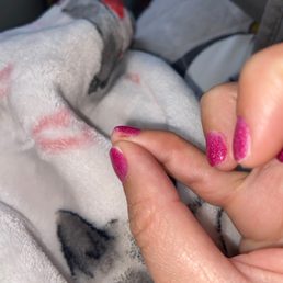 The image shows a hand with pink glitter nail polish holding a person's finger. The background appears to be a towel or blanket with a soft, textured surface and some printed or patterned areas. The focus is on the hand and nails, with the background slightly blurred.