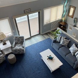 A living room features a blue carpet, a gray sofa with patterned pillows, an armchair with a coastal pattern, and decorative items including a turtle figurine on the coffee table.