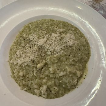 Peacock Cafe - Crab and Pesto Risotto - A white plate contains crab and pesto risotto topped with grated cheese, served at a restaurant. A glass of beverage is visible in the background. - Wine Bars Near Me - Washington, DC, District of Columbia