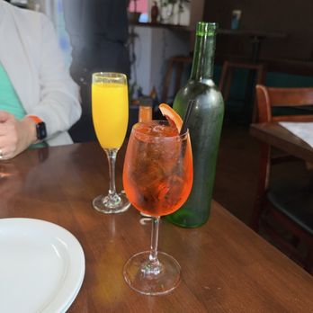 FOB Kitchen - Aperol spritz and a mimosa - The image shows three beverages on a wooden table in a restaurant. The red cocktail is in a tall glass with ice, garnished with an orange slice and a straw. Behind it, a yellow mimosa is in a flute glass. A green bottle is also present on the table. Part of a person wearing a white jacket and a turquoise shirt is visible on the left, and a person in a dark jacket is standing in the background. The restaurant interior includes wooden chairs, shelves, and some greenery. - Filipino Near Me - Oakland, California