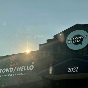Beyond Hello - Front of the building - The image depicts the exterior of Beyond Hello, a cannabis dispensary, with visible signage including the business name and description. The building features a modern facade with the address 2021 displayed prominently. The sky is clear, and the sun is partially visible, creating lens flare effects in the photo. - Cannabis Clinics Near Me - Sauget, Illinois