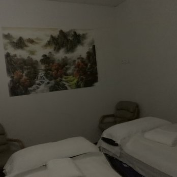 Luna Spa - Room - The image shows a hotel room with minimal lighting, containing two beds with white bedding, each with folded towels on top. There are two chairs positioned against the walls, and a landscape painting depicting mountains, trees, and waterfalls hangs on the wall behind the beds. - Massage Near Me - Plainfield, Indiana