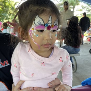 A Face Painter - Unicorn face paint - A girl with elaborate unicorn face paint, including flowers and rhinestones, is seated outdoors under a pavilion. - Face Painting Near Me