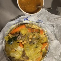 Lamb Couscous - The image shows a takeout meal with a round aluminum container filled with vegetable couscous, including carrots, zucchini, chickpeas, cauliflower, and leafy greens. A cup of soup with herbs and a spoon is placed above the container. The meal is on a gray surface with a plastic bag and napkin nearby.