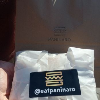 Paninaro - A person holds a wrapped food item with a black sticker showing @eatpaninaro, in front of a Paninaro paper bag, likely from a restaurant in Ottawa. - Sandwiches Near Me - Ottawa, Ontario