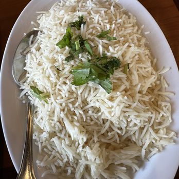 Curry Masala - Spring - Plain Basmati Rice - The image shows a white oval ceramic plate filled with plain cooked basmati rice, garnished with fresh cilantro leaves on top. A metal spoon rests on the left side of the plate. The plate is placed on a wooden surface, and part of a glass is visible at the top of the image. - Indian Near Me - Spring, Texas