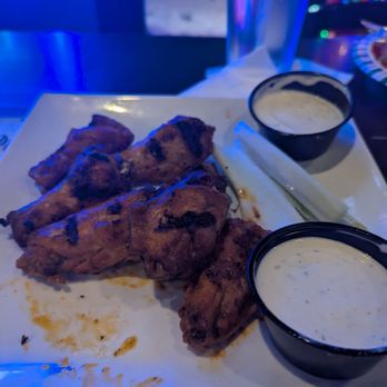 Trail Stop Tavern - Buffalo wings - Plate of buffalo wings with visible grill marks and sauce, accompanied by two cups of creamy dipping sauces, likely ranch or blue cheese, on a white square plate. A glass of water and a napkin are also present, with colorful holiday lights visible in the background, indicating a lively dining environment at Trail Stop Tavern in Eagan, MN. - American Near Me - Eagan, Minnesota