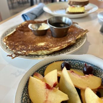 Borage - Seasonal fruit side, pancake, breakfast sandwich - The image shows a breakfast scene with a plate of a large crepe topped with butter and syrup, accompanied by two small metal containers of syrup and butter. In the foreground, there is a bowl filled with sliced peaches and grapes. In the background, a plate with a breakfast sandwich, including an egg, bacon, and bread, is visible. The setting appears to be in a casual dining environment with chairs and tables visible in the background. - Breakfast & Brunch Near Me - Indianapolis, Indiana