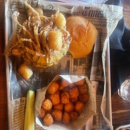 Sweet Potato Tater Tots Burger - A tray lined with newspaper holds a basket of golden-brown tater tots, a bread roll, a pickle spear, and a serving of fried seafood mix with onion rings and other battered items. The tray is on a wooden table with black napkins and a glass of water nearby.