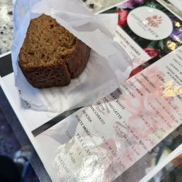 Pumpkin Loaf - A slice of pumpkin loaf with a moist, dense texture is placed on a white paper wrapper. The loaf is positioned on top of a menu featuring various coffee and tea options, including lattes, mochas, and infusions. The menu has a floral background and is partially visible, with prices listed for each beverage. The setting appears to be a coffee shop or café, with a granite countertop and some blurred objects in the background.