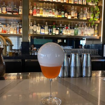 Verdigris - Dragon's Folly - A cocktail featuring a sizable foam head is positioned on a marble bar counter. The background includes shelves filled with various liquor bottles, bar tools, and a few patrons visible through a mirror. The setting appears to be an upscale bar or cocktail lounge. - Tapas/Small Plates Near Me - Leawood, Kansas