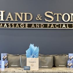 Hand & Stone Massage and Facial Spa - "Hand & Stone Massage and Facial Spa" sign in gold letters on gray wall. Display of blue Dermalogica product boxes, gray Derm...