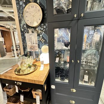 IKEA - Glassware and tableware are displayed in a dark cabinet and on a small table, with a decorative floral wall clock in the background. - Kitchen & Bath Near Me - Brooklyn, New York