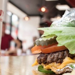 A close-up photograph of a Five Guys cheeseburger with pickles, tomatoes and jalapenos in a lettuce wrap.