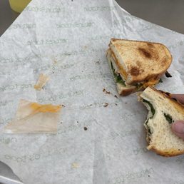 They left the cheese paper in the sandwich. The sandwich needed a sauce and I asked for one but they said no. I want my dollar tip back. - A sandwich with toasted bread, visible greens, and cheese, partially eaten, on branded paper with crumbs and cheese remnants.