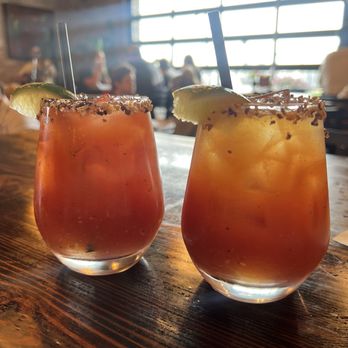 Yorgo's Bageldashery - Bloody Mary - Two Bloody Mary drinks with lime wedges and salted rims are placed on a wooden table inside a restaurant with large windows and blurred patrons in the background. - Bagels Near Me - Norfolk, Virginia