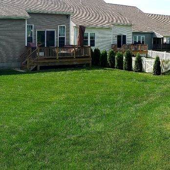Egan Landscape Group - The view of our back yard, before the patio. - The image shows a backyard with a neatly mowed green lawn extending across the scene. On the left, there are neighboring houses with decks and windows, and a white fence separates the yards. On the right side, there are trees and shrubs, with a garden lamp visible among the greenery. The scene appears to be in a suburban residential area. - Landscaping Near Me - Plymouth, Massachusetts