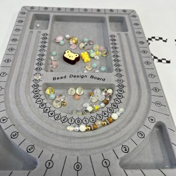 Crafts Zone Studio - A bead design board labeled 'Bead Design Board' contains an assortment of small beads, including pastel-colored, metallic, and clear beads, along with a bear-shaped charm and other decorative elements. The board has numbered sections and a central area for arranging beads during jewelry or craft projects. - Paint & Sip Near Me - Brookline, Massachusetts