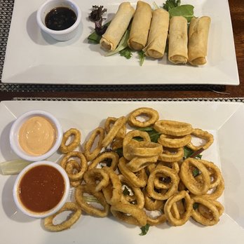 Connolly’s Corner - Vegetable Spring Rolls Crispy Fried Calamari - A photo shows two rectangular white plates with Asian appetizers: spring rolls with soy sauce and fried calamari with dipping sauces, on a woven placemat. - Sports Bars Near Me - Maspeth, New York