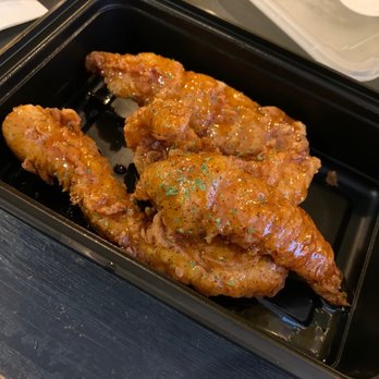 Filly Luv - Hot honey tenders - Three glazed chicken tenders coated in sauce and herbs are arranged in a black takeout container. The tenders are seasoned and appear crispy, with visible herbs on top. The container is on a dark surface, and a clear plastic lid is partially visible in the background. - Chicken Shop Near Me - Brooklyn, New York