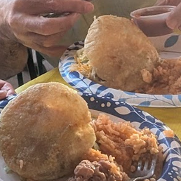 Gorditas - Two plates of gorditas, a traditional Mexican dish, are held by hands in the image. Each plate contains a large, round, fried gordita with sides of rice, beans, and possibly meat. A small container of green sauce is visible in the background, along with a person holding a drink. The setting appears to be a casual dining environment.