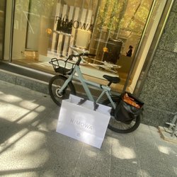 Rimowa - A blue vanmoof electric bike in front of the beautiful golden rimowa store window display. In front of it sits a large rimowa shopping bag.