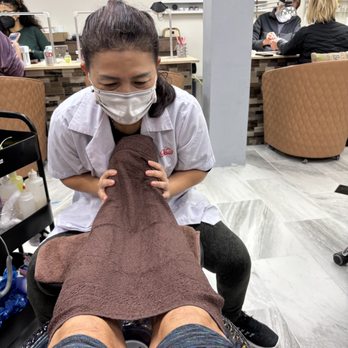 Fantasy Nails - A woman wearing a face mask and a white uniform is receiving a pedicure at a nail salon. She is seated with her legs extended, covered with a brown towel. The salon interior includes nail polish displays on the wall, a manicure station in the background, and other clients and staff. The environment appears clean and organized, with multiple clients and staff members visible. - Nail Salons Near Me - Eugene, Oregon