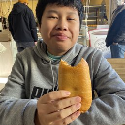 Sausage Kolaches - A young person with short black hair, wearing a gray hoodie, is seated at a wooden table in a cafe. They are holding a large pastry with a bite taken out of it, and are looking at the camera with a slight smile. In the background, there are balloons, a person wearing a yellow beanie and black jacket, and another person with a face mask, indicating a casual indoor setting.