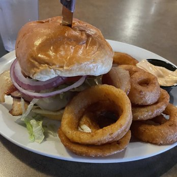 Dawsons On Main - Grilled Chicken Club Sandwich with onion rings - A chicken sandwich with a toasted bun, lettuce, tomato, onion slices, and grilled chicken, accompanied by a pile of onion rings and a small container of dipping sauce on a white plate. The plate is placed on a wooden table in a restaurant setting, with a glass of water visible in the background. - American Near Me - Indianapolis, Indiana