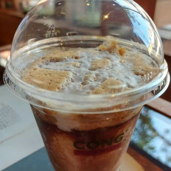 Cong Caphe - White Coffee - A cold coffee drink with whipped cream topping is served in a clear plastic cup with a dome lid, placed on a table inside a cafe. - Coffee & Tea Near Me - Toronto, Ontario
